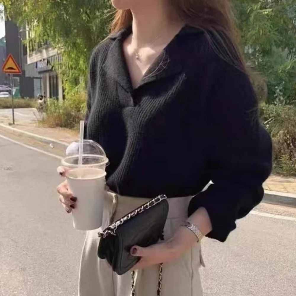 Frau in schwarzem Pullover hält Eiskaffee und schwarze Handtasche, steht auf sonniger Straße. Mode, Streetstyle, Herbstoutfit, urbane Kulisse.