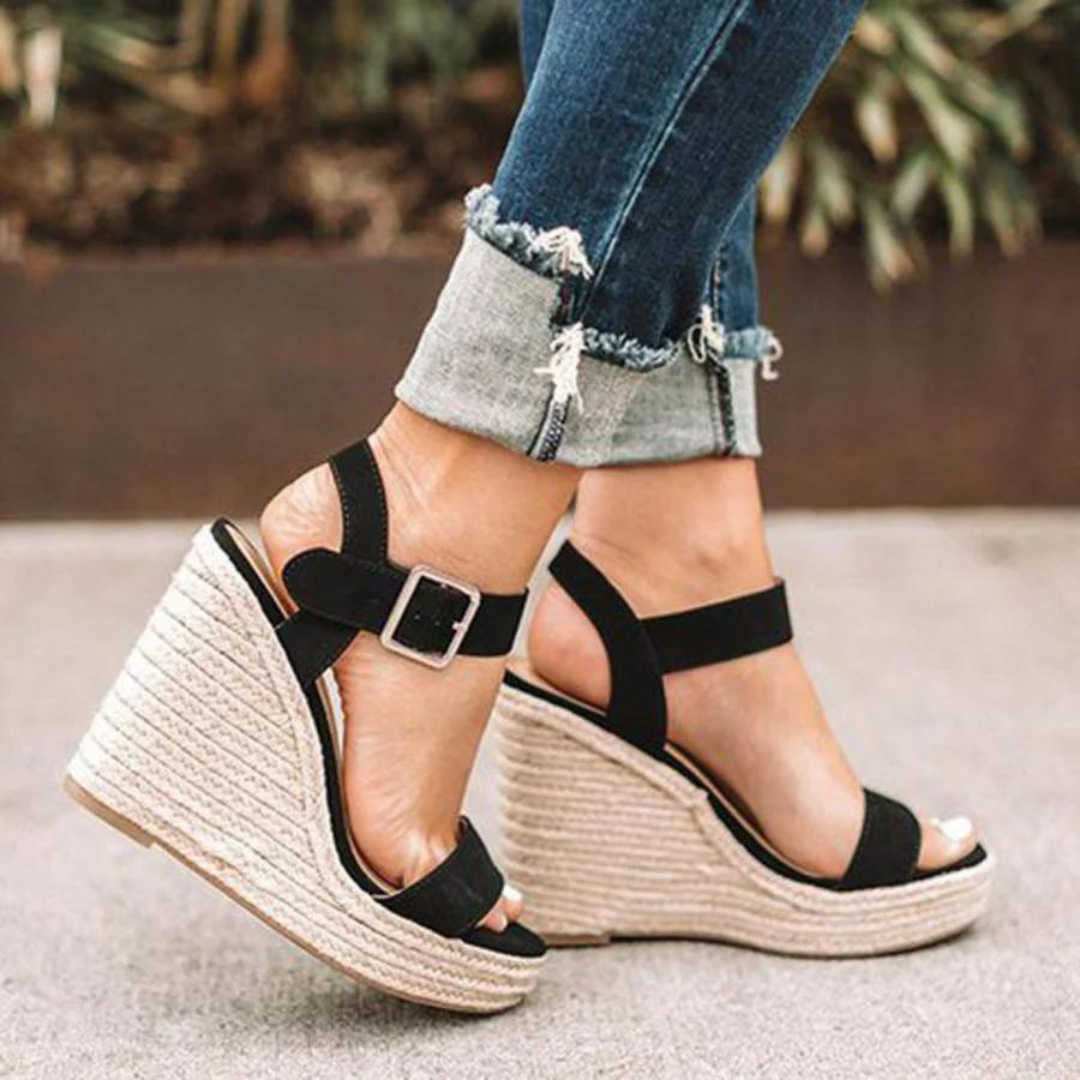 Frau trägt schwarze Keilsandaletten mit Bastsohle und Schnalle, kombiniert mit hochgekrempelten Jeans. Modische Sommerschuhe, Damenmode.