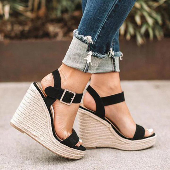 Frau trägt schwarze Keilsandaletten mit Bastsohle und Schnalle, kombiniert mit hochgekrempelten Jeans. Modische Sommerschuhe, Damenmode.