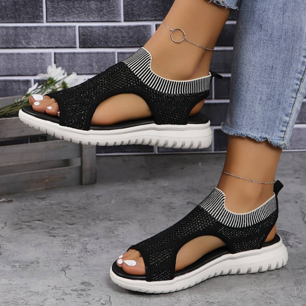 Schwarze Damen-Sandalen mit weißer Sohle, gestricktem Obermaterial und offener Zehenpartie, getragen mit blauen Jeans vor einer Ziegelwand.