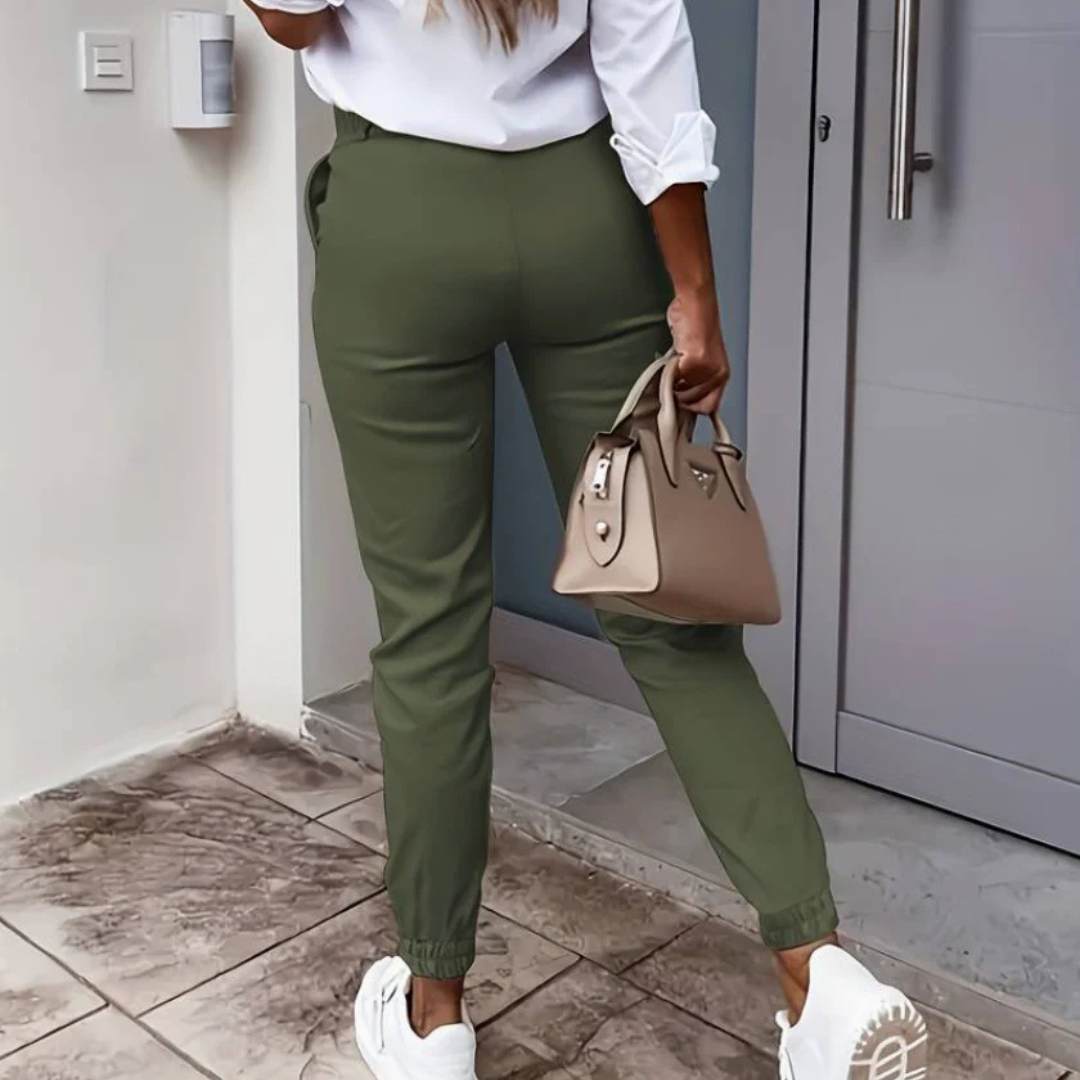 Frau in grüner Hose und weißem Hemd, trägt beige Handtasche und weiße Sneaker, steht vor moderner Haustür. Mode, Streetstyle, Damenbekleidung.