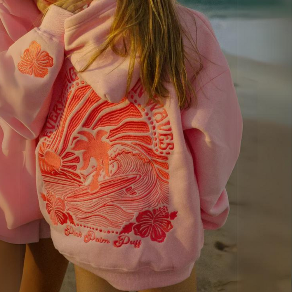 Mädchen in rosa Hoodie mit Surfmuster und Hibiskusblüten am Strand bei Sonnenuntergang. Strandmode, Kinderbekleidung, Surfer-Style.