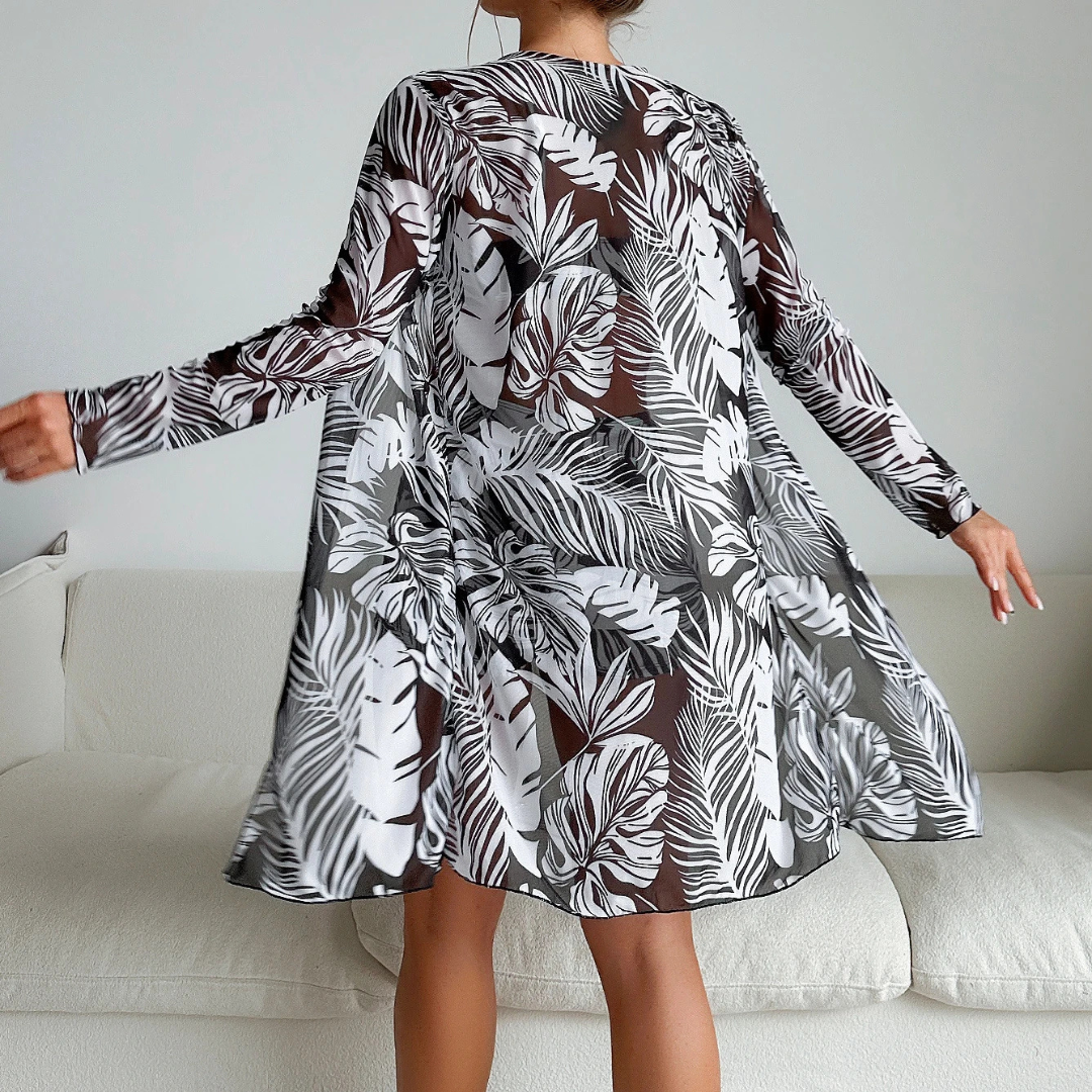 Frau in geblümtem, transparentem Kleid mit langen Ärmeln, steht vor einem weißen Sofa. Tropisches Muster, modisch, elegant, modern.