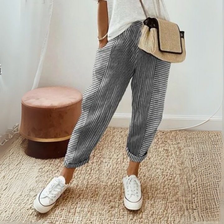 Frau in gestreifter Hose und weißem T-Shirt, trägt eine Strohtasche und weiße Sneaker, steht auf Teppich neben rosa Hocker. Modetrend, lässiger Stil.