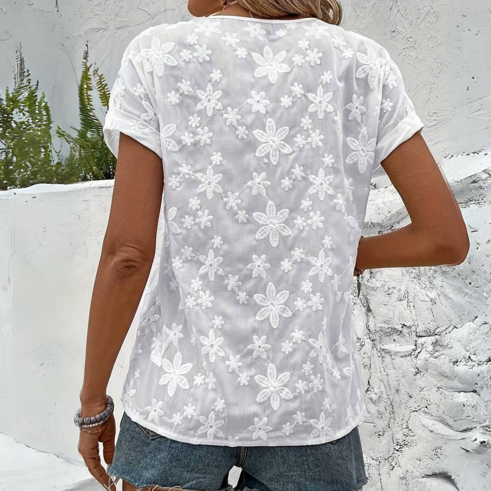 Frau in weißem, transparentem Spitzen-T-Shirt mit Blumenmuster, trägt Jeansshorts, steht vor weißer Wand. Sommermode, lässiger Stil.