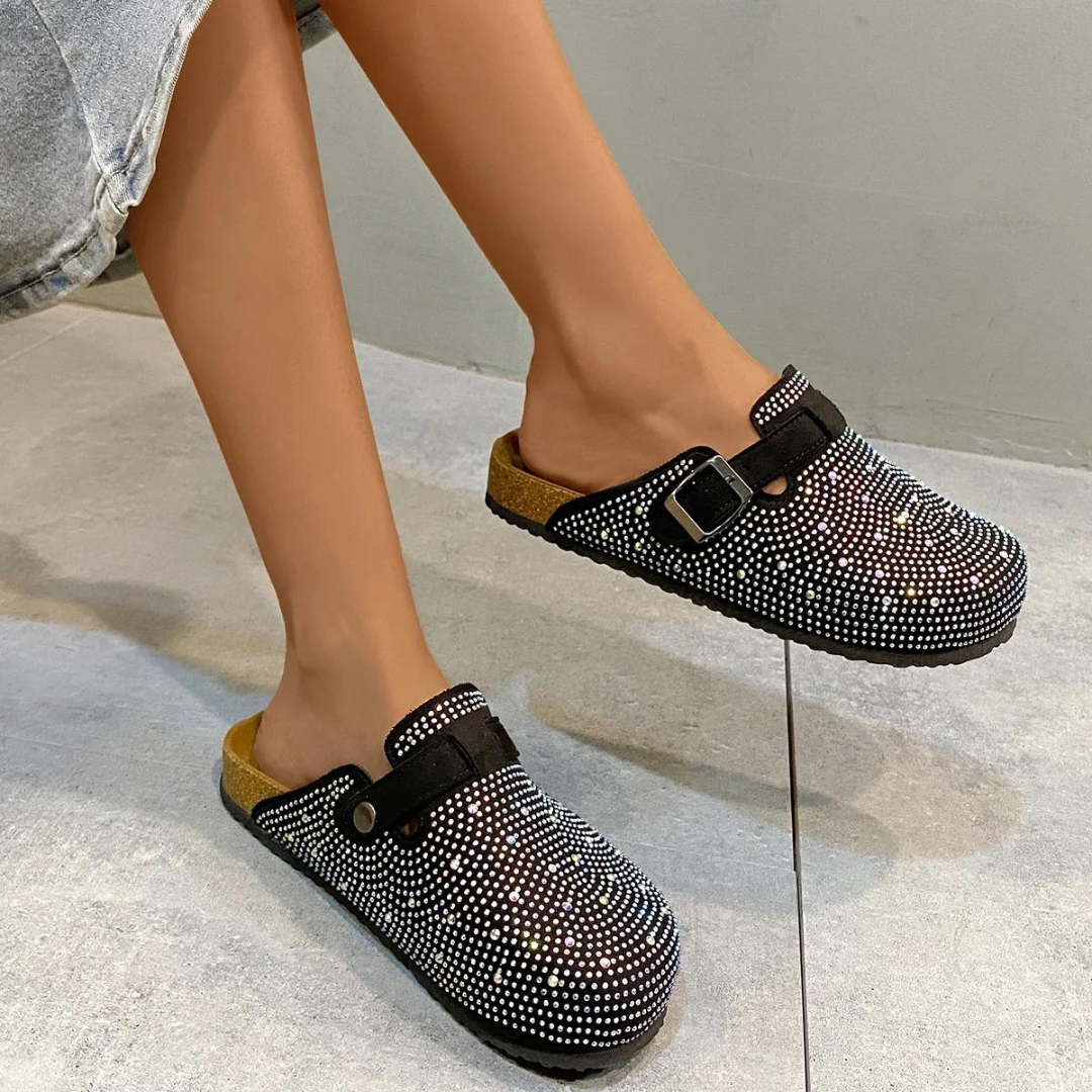 Damenfüße in modischen, schwarzen Clogs mit Strasssteinen und Schnalle auf grauem Boden. Trendige Schuhe, bequem, stilvoll.
