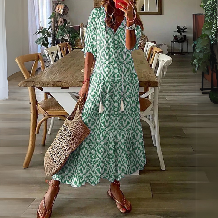 Frau in grünem Boho-Kleid mit Ethno-Muster, hält eine geflochtene Tasche, steht in modernem Esszimmer mit Holztisch und Pflanzen.