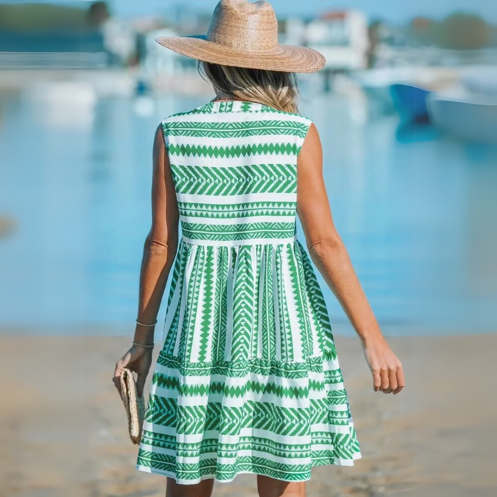 Frau in grün-weißem Sommerkleid und Strohhut am Strand, hält eine Clutch. Perfektes Outfit für Sommermode und Strandurlaub.