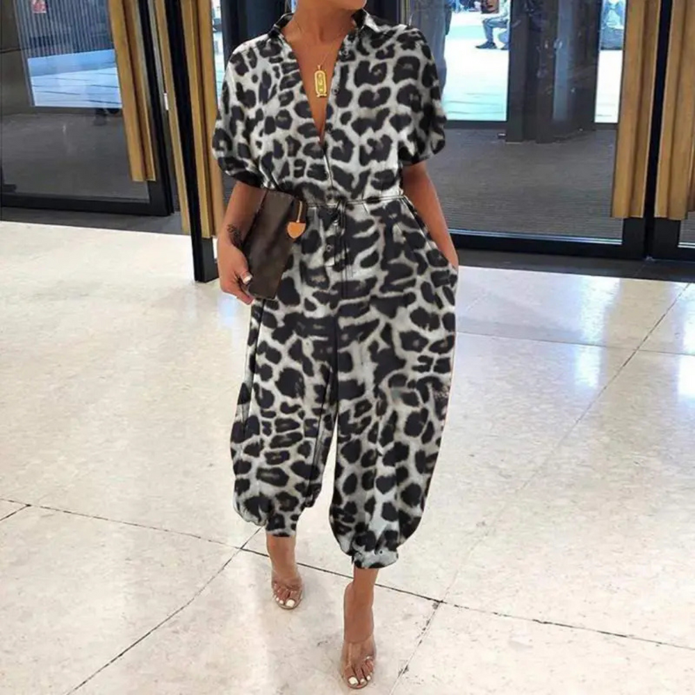 Frau in modischem Leopardenmuster-Jumpsuit, steht in modernem Innenraum. Trendige Damenmode, stilvoller Einteiler, elegantes Design.