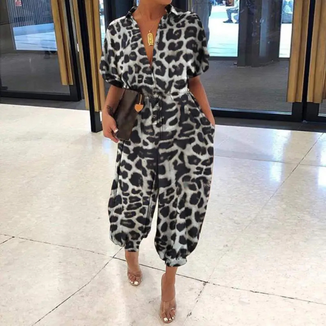 Frau in modischem Leopardenmuster-Jumpsuit, steht in modernem Innenraum. Trendige Damenmode, stilvoller Einteiler, elegantes Design.
