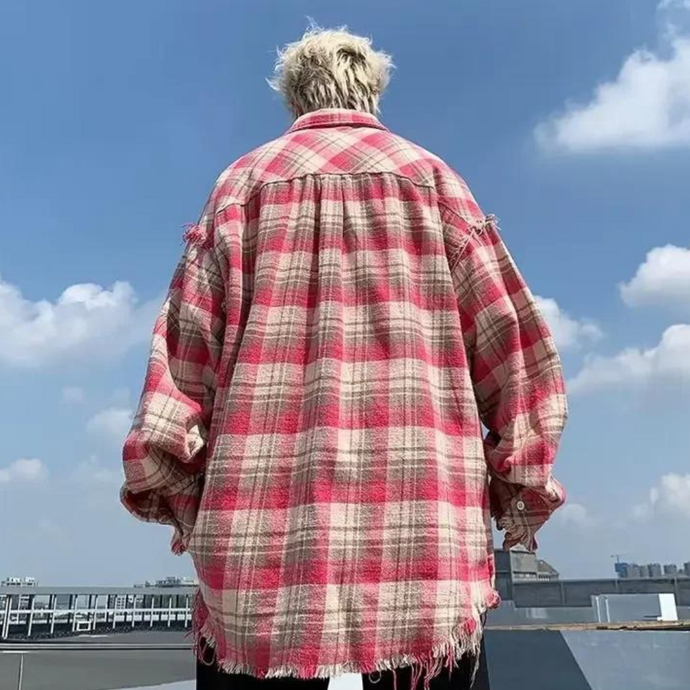 Rückansicht einer Person in übergroßem, rot-kariertem Flanellhemd mit Fransen, stehend auf einem Dach vor blauem Himmel. Urbaner Streetwear-Stil.