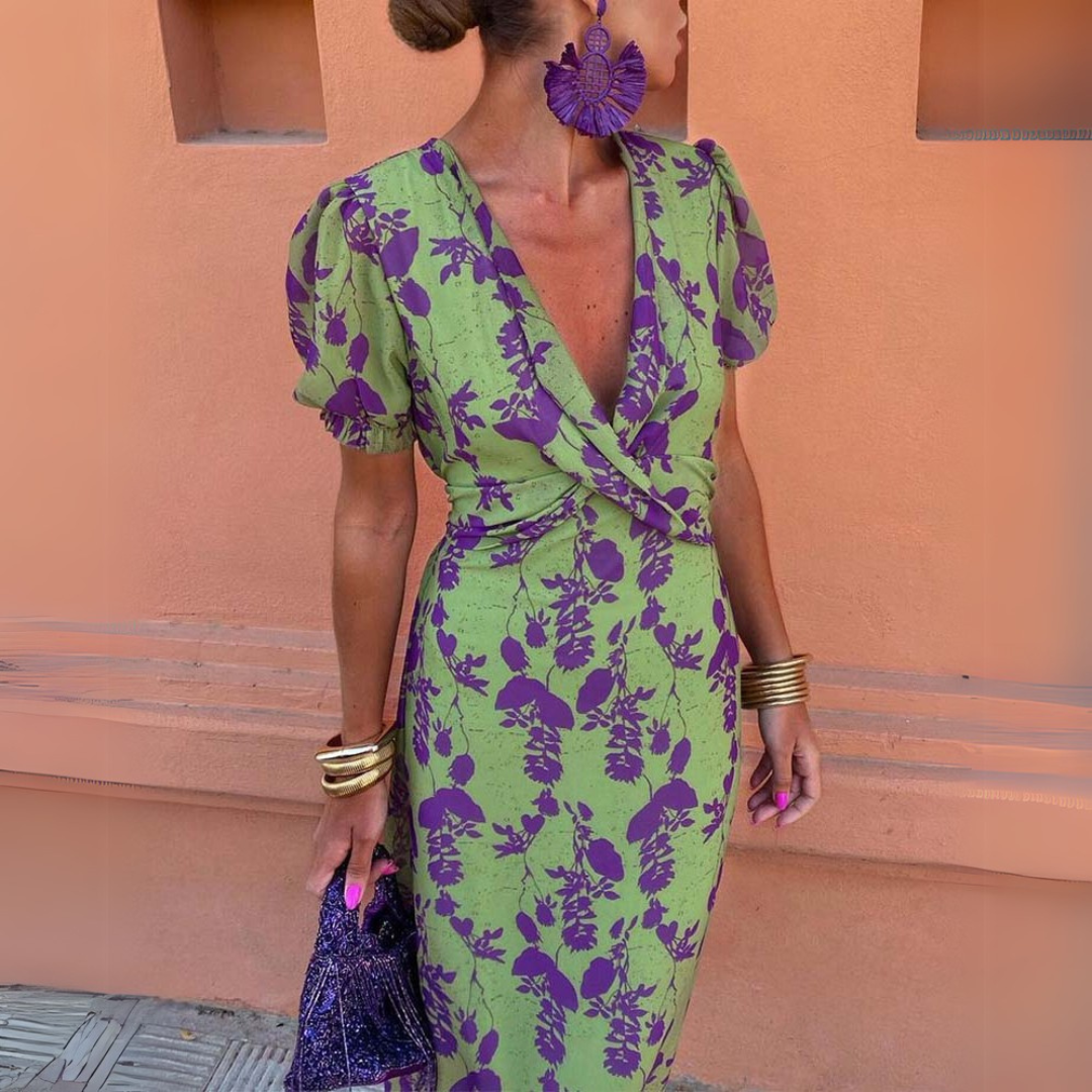 Frau in grün-lila geblümtem Kleid mit Puffärmeln, großen lila Ohrringen und goldenen Armreifen vor orangefarbener Wand. Elegante Mode, Sommerkleid.