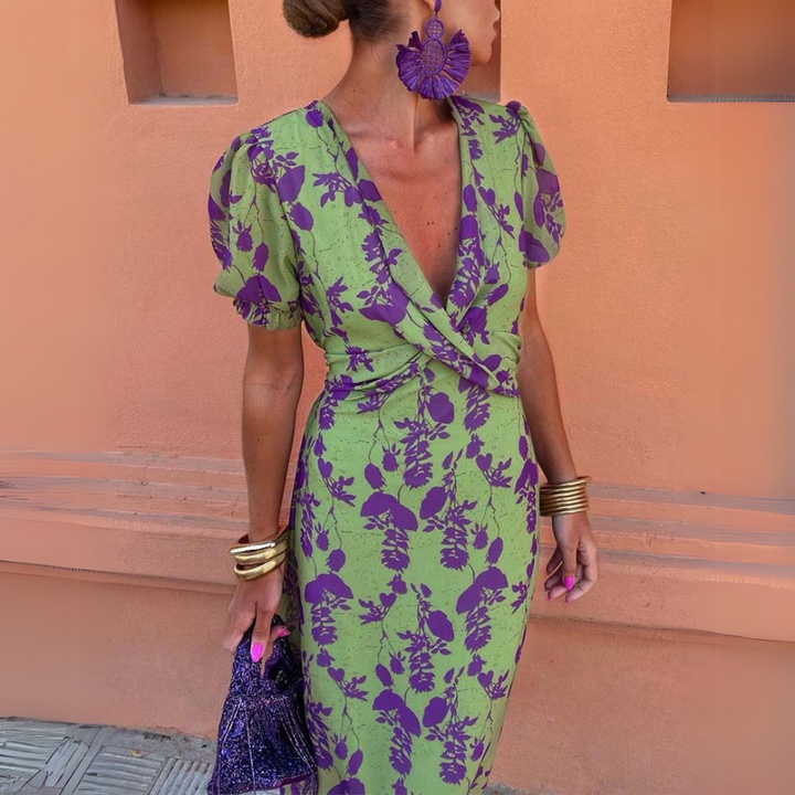 Frau in grün-lila geblümtem Kleid mit Puffärmeln, großen lila Ohrringen und goldenen Armreifen vor orangefarbener Wand. Elegante Mode, Sommerkleid.