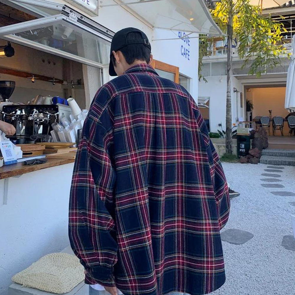 Mann in kariertem Hemd und schwarzer Kappe steht vor einem Café im Freien. Urbaner Stil, Freizeitkleidung, entspannte Atmosphäre.