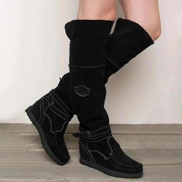 Schwarze Damenstiefel aus Wildleder, kniehoch mit Schnallen und Ziernähten, auf Holzboden. Modische Herbst-Winter-Schuhe für Frauen.