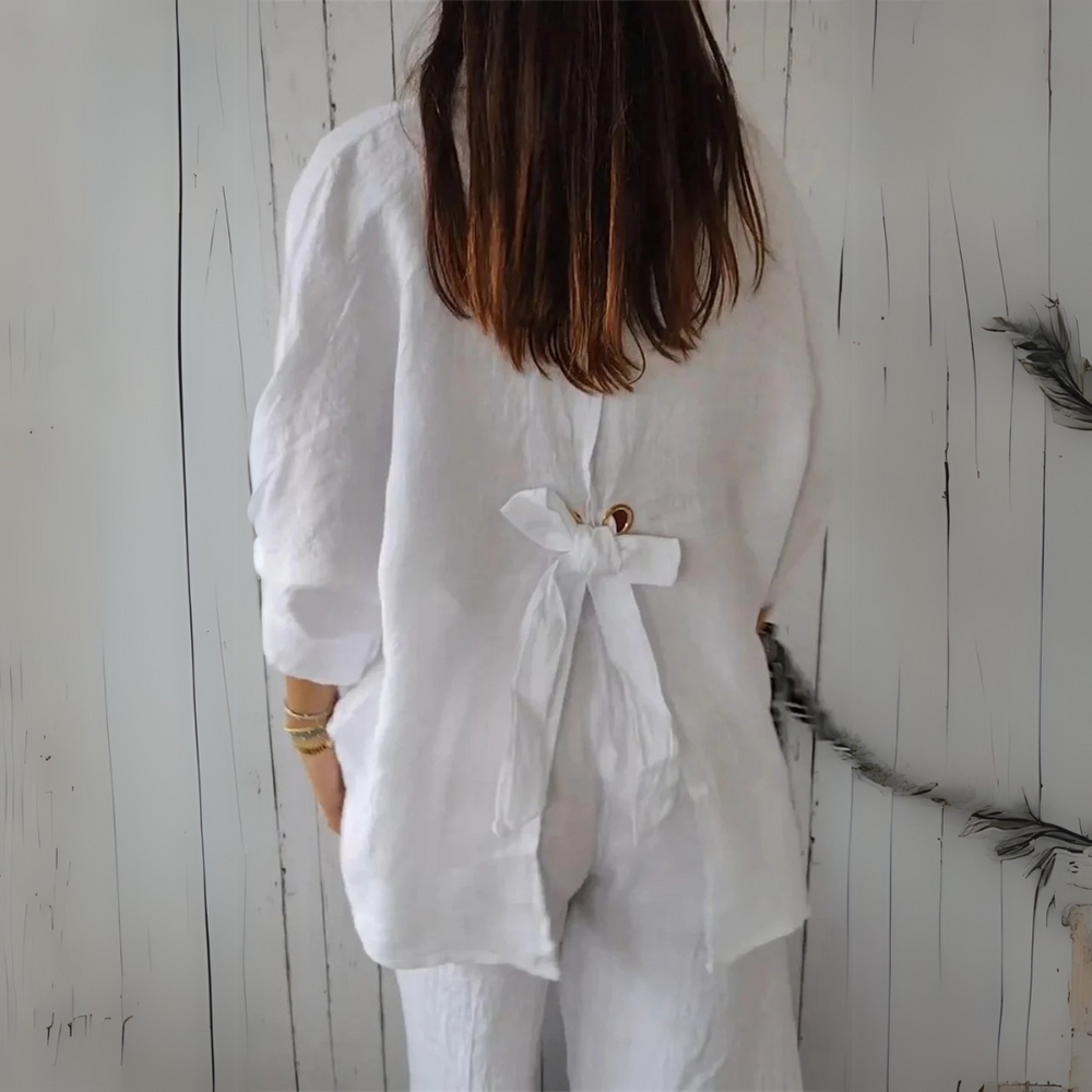 Frau in weißem Leinen-Outfit mit Schleife am Rücken, vor rustikalem Holzhintergrund. Modetrend, nachhaltige Kleidung, Sommerlook.
