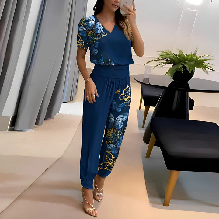 Frau in blauem, geblümtem Jumpsuit posiert vor Spiegel. Elegante Abendmode, modische Kleidung, stilvolle Damenbekleidung, Innenraum mit Pflanzen.
