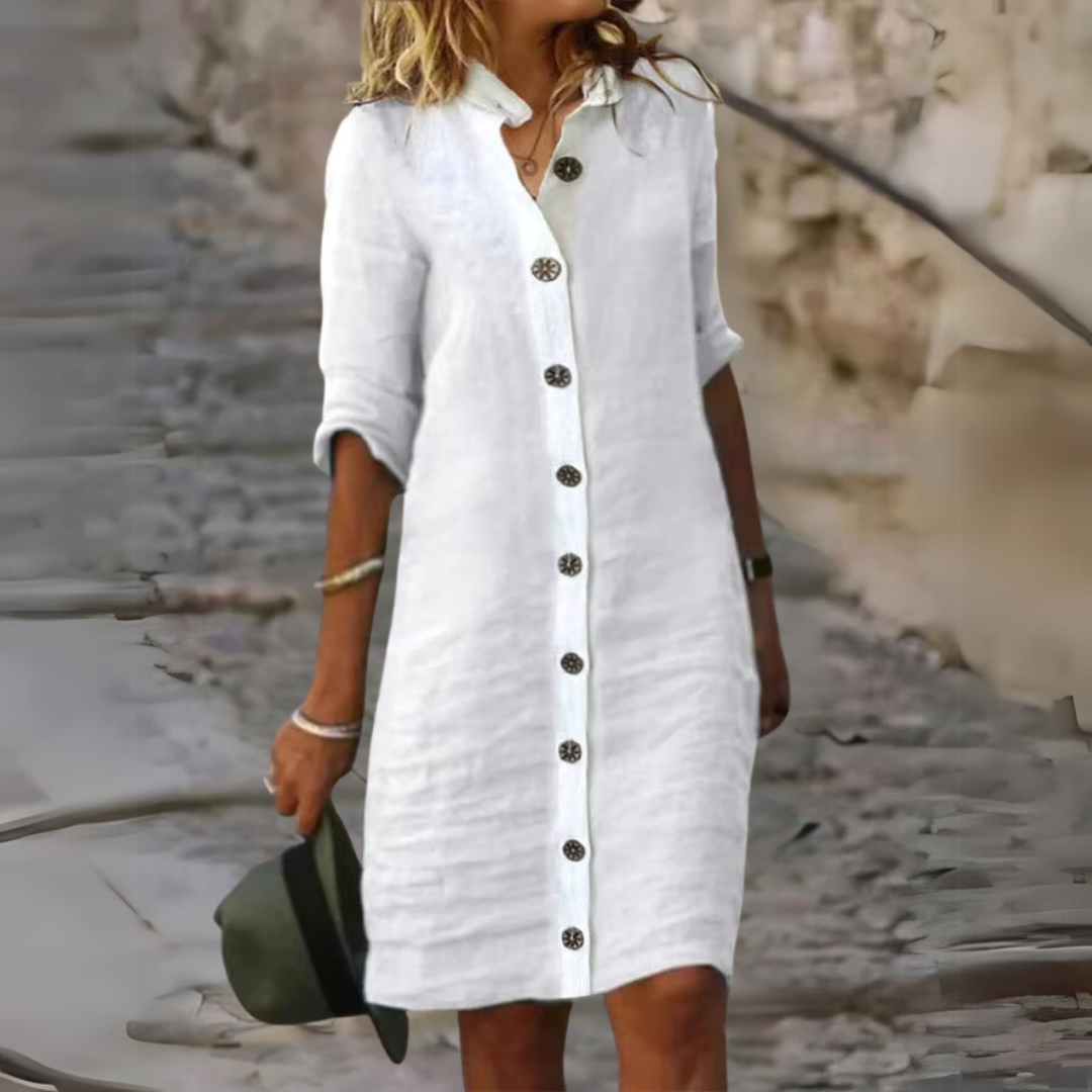 Frau in weißem Leinenkleid mit Knopfleiste, lässig auf Kopfsteinpflaster gehend, hält Hut. Sommermode, Freizeitkleidung, stilvoll und elegant.