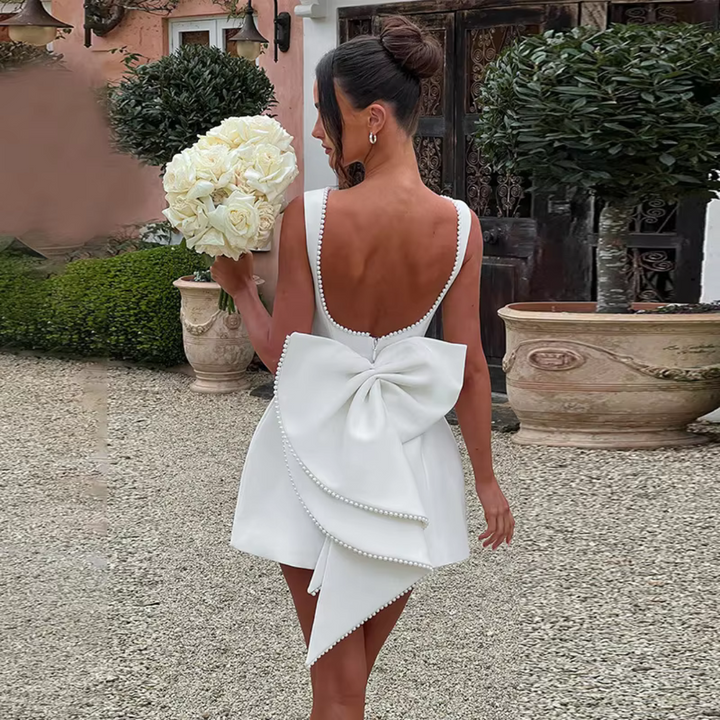 Frau im eleganten weißen Kleid mit großer Schleife, hält Blumenstrauß. Hochzeit, Brautmode, stilvolle Gartenkulisse.