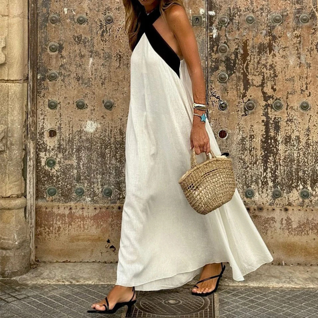 Frau in weißem Maxikleid mit schwarzem Träger, hält geflochtene Strohtasche, steht vor antiker Holztür. Sommermode, Boho-Stil, Stadtbummel.