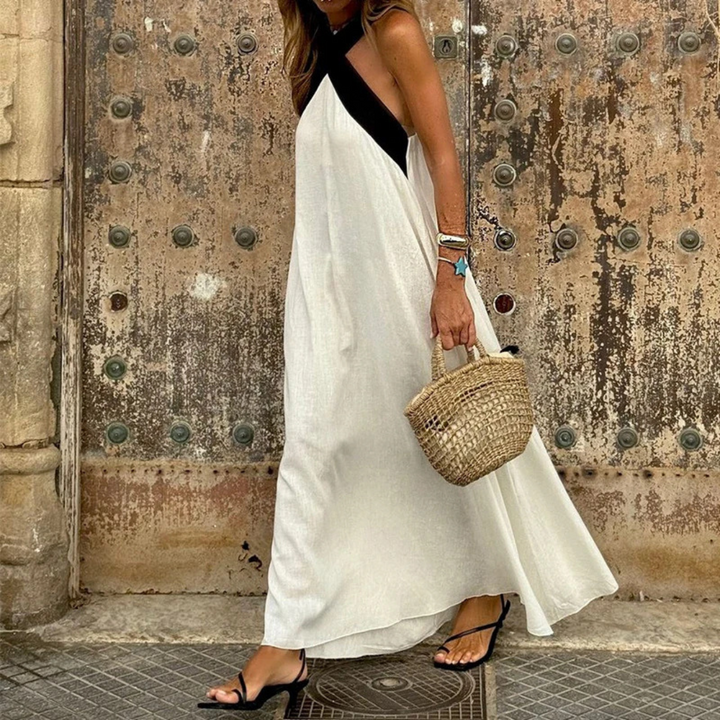 Frau in weißem Maxikleid mit schwarzem Träger, hält geflochtene Strohtasche, steht vor antiker Holztür. Sommermode, Boho-Stil, Stadtbummel.