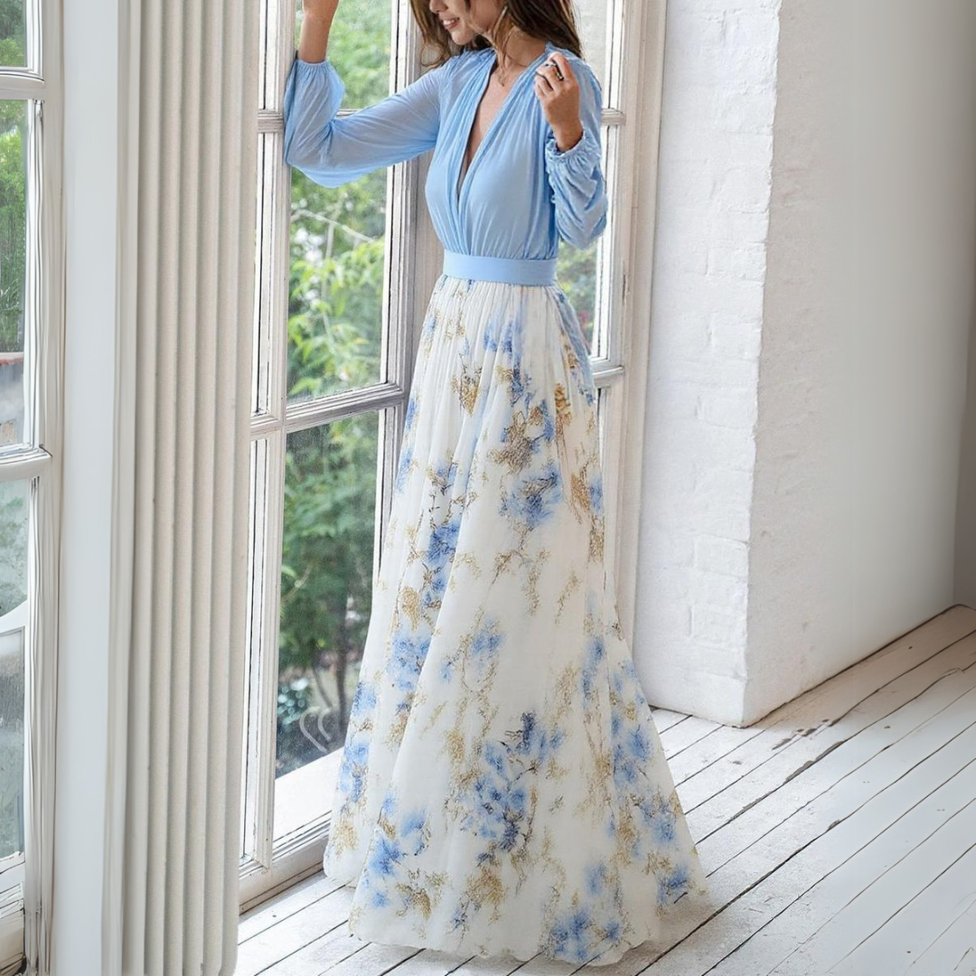 Frau in elegantem, langem Kleid mit blauem Oberteil und floralem Rock steht am Fenster. Stilvolle Abendmode, modisches Design, Sommerkleid.