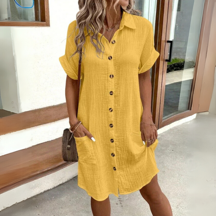 Blonde Frau in gelbem Leinenhemdkleid mit Knopfleiste und Taschen, steht vor einem modernen Gebäude. Sommermode, lässiger Stil, Damenbekleidung.