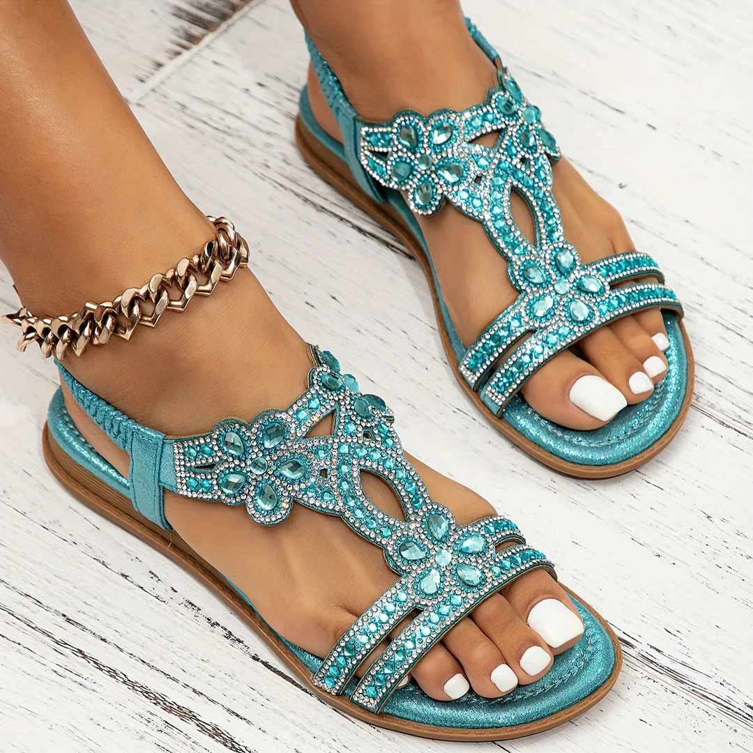 Türkise Sandalen mit Strasssteinen und Blumenmuster auf weißem Holzboden, getragen von einer Person mit goldener Fußkette und weiß lackierten Nägeln.