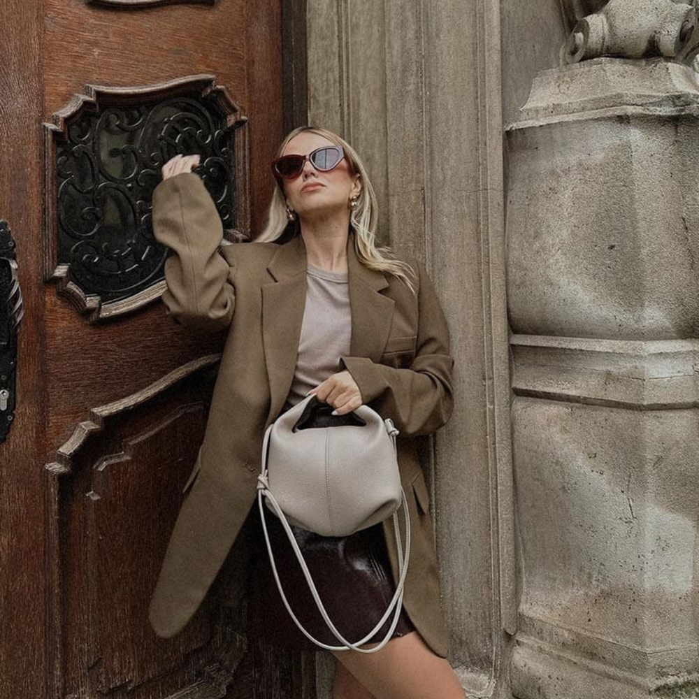 Frau in braunem Blazer und Sonnenbrille lehnt an Holztür, hält graue Handtasche. Mode, Streetstyle, Herbstlook, urbane Eleganz.