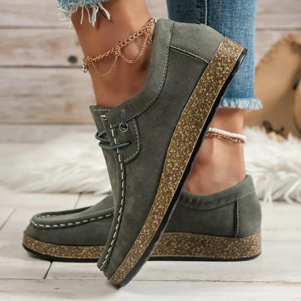 Graue Damen-Mokassins mit Korksohle, getragen mit Jeans und goldenen Fußkettchen, auf Holzboden. Bequeme Freizeitschuhe, modischer Look.