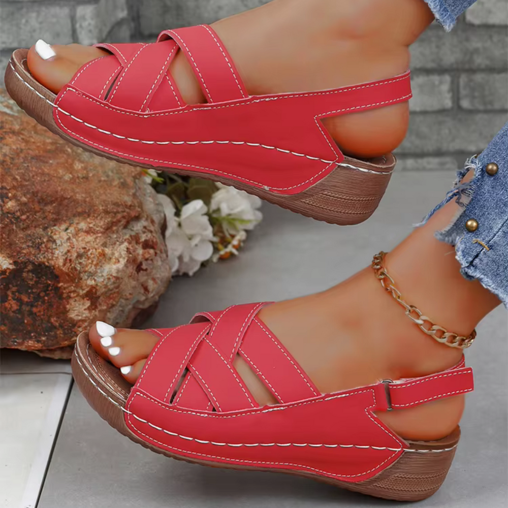 Rote Sandalen mit Keilabsatz, Kreuzriemen und weißer Naht, getragen von einer Person mit Jeans und Fußkettchen, vor einem Stein und Blumen.
