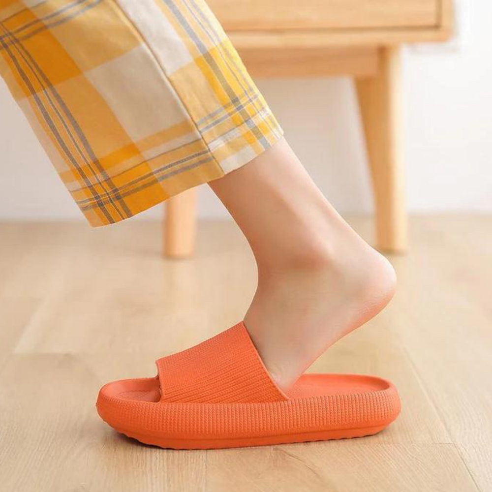 Orange Hausschuhe auf Holzfußboden, getragen von Person in karierten gelben Hosen. Bequeme, modische Pantoffeln für Zuhause.