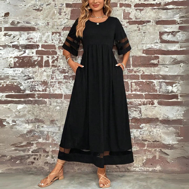 Frau in elegantem, schwarzem Maxikleid mit transparenten Ärmeln vor einer Backsteinwand. Modetrend, Abendkleid, Damenmode, stilvoll.