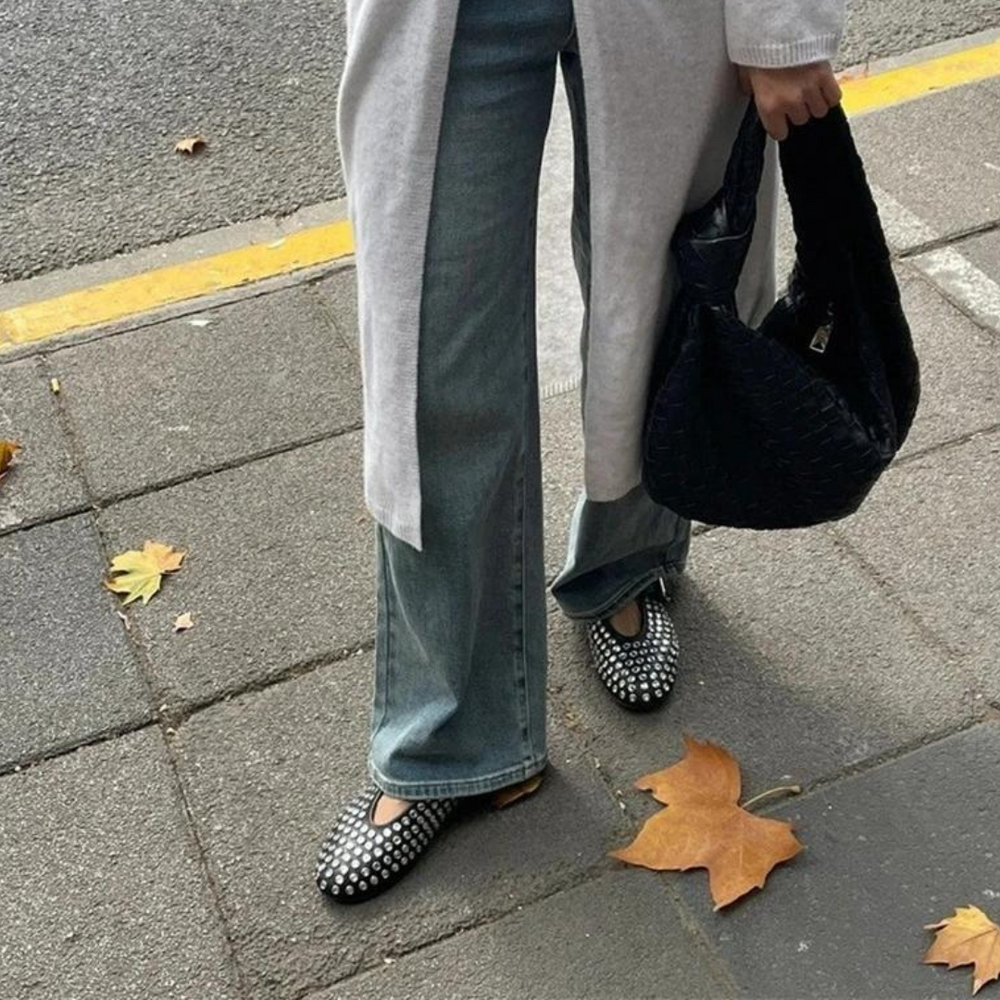 Frau mit blauer Jeans und karierten Ballerinas auf Gehweg, hält schwarze Tasche. Herbstlaub auf Boden. Straßenmode, lässiger Stil.