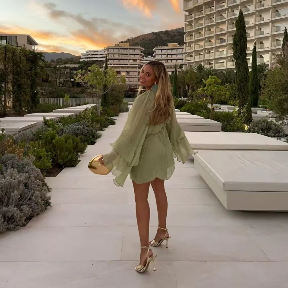 Eine Frau in einem grünen Kleid und High Heels geht auf einer Terrasse bei Sonnenuntergang. Im Hintergrund moderne Gebäude und Bäume.