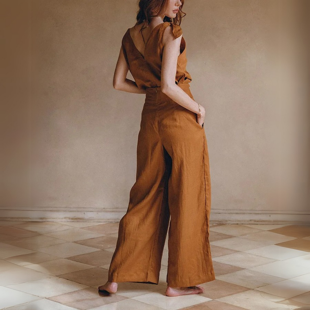 Frau in braunem Leinen-Jumpsuit, barfuß auf gemustertem Boden. Modetrend, nachhaltige Kleidung, eleganter Einteiler, Rückansicht.