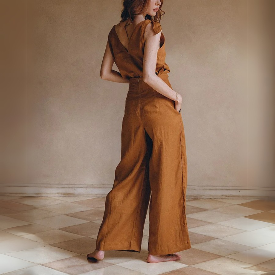 Frau in braunem Leinen-Jumpsuit, barfuß auf gemustertem Boden. Modetrend, nachhaltige Kleidung, eleganter Einteiler, Rückansicht.