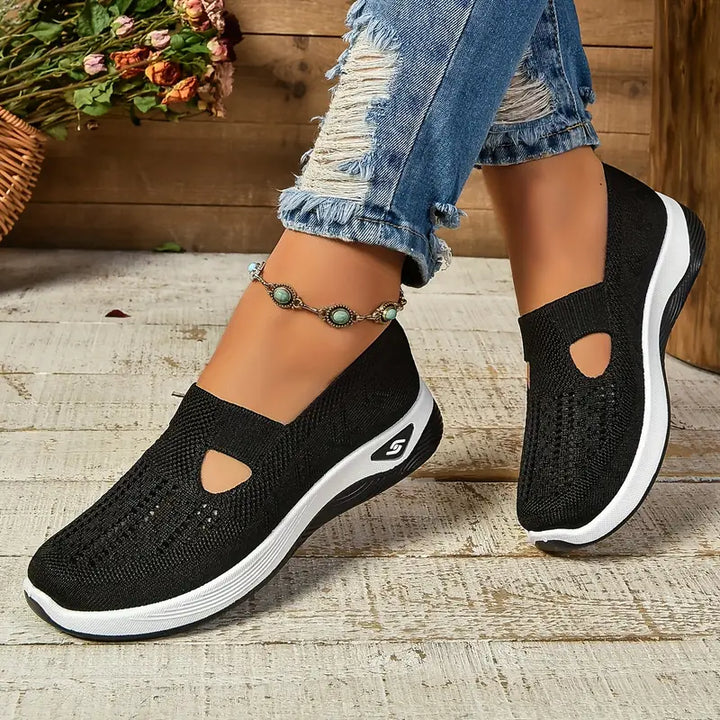 Schwarze Damen-Sneaker mit Keilabsatz, atmungsaktivem Strickmaterial und Cut-Out-Design, kombiniert mit zerrissenen Jeans auf Holzboden.