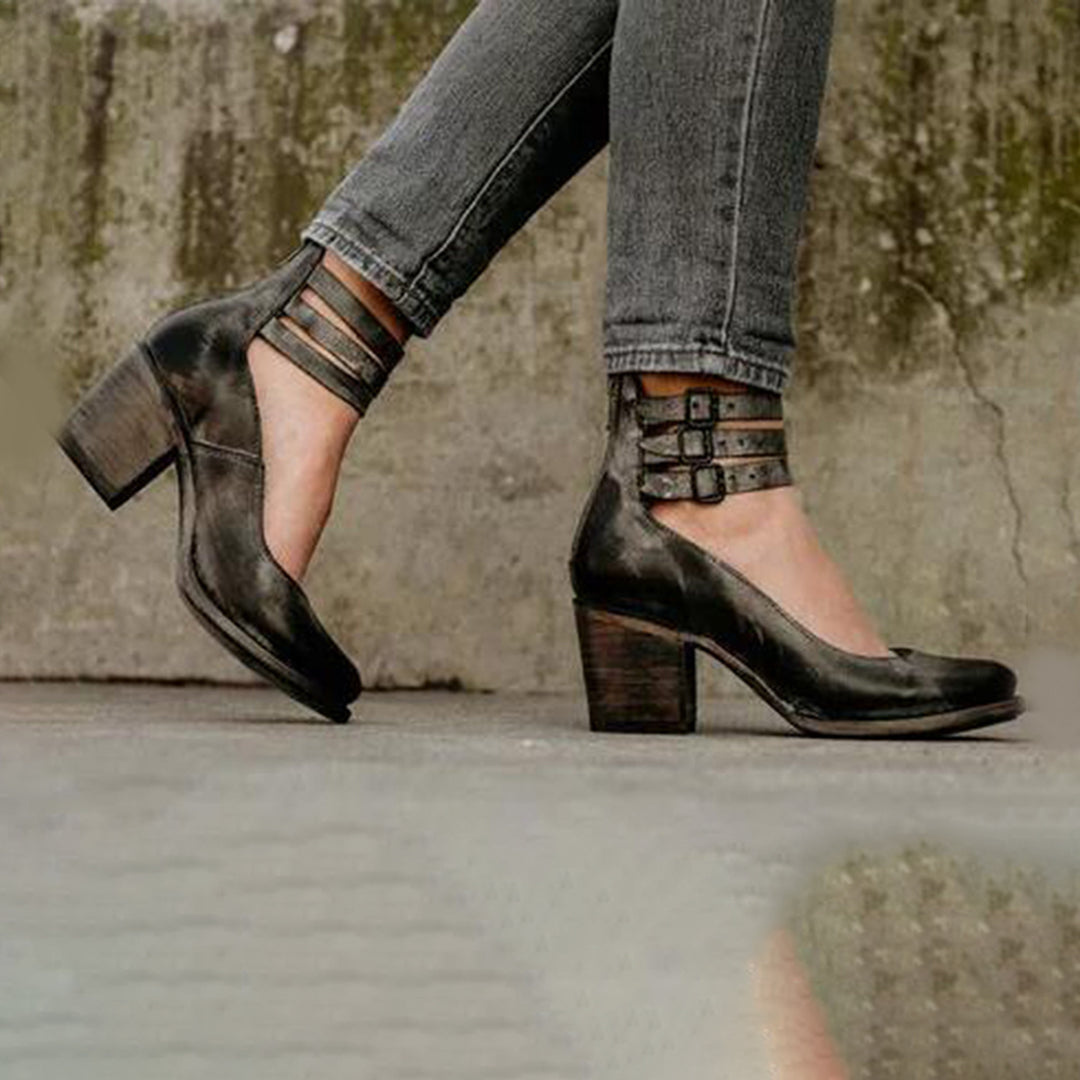 Damen schwarze Lederpumps mit Blockabsatz und Riemchen, getragen zu grauen Jeans, vor einer Betonwand. Modische Schuhe, elegantes Design.