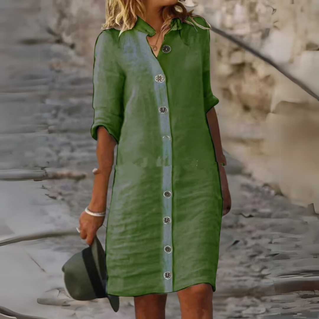Frau in grünem Leinenkleid mit Knopfleiste, hält Hut in der Hand, steht auf gepflastertem Weg. Sommermode, Freizeitkleidung, stilvoll.