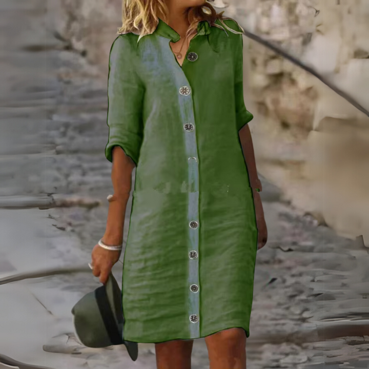 Frau in grünem Leinenkleid mit Knopfleiste, hält Hut in der Hand, steht auf gepflastertem Weg. Sommermode, Freizeitkleidung, stilvoll.