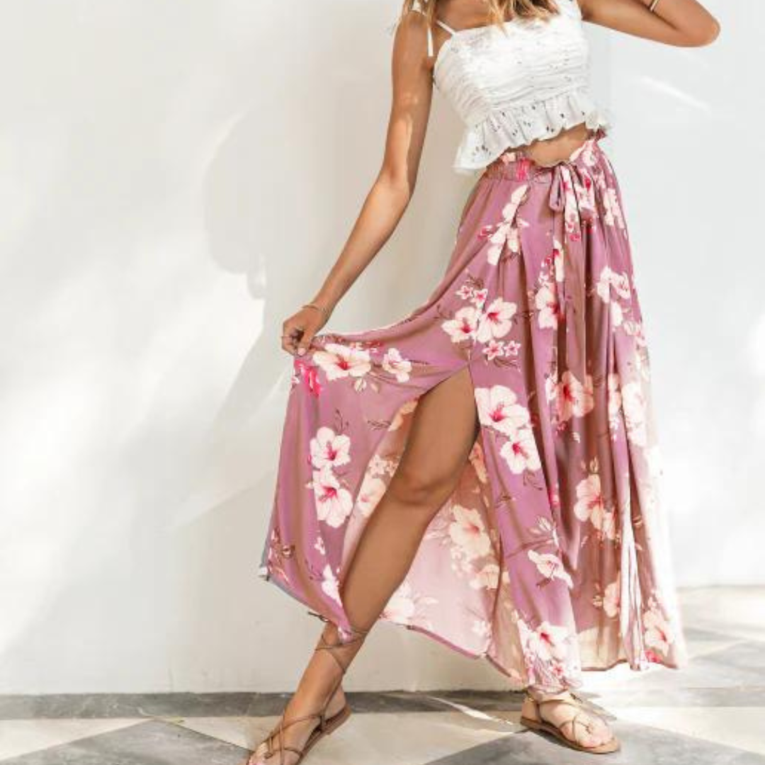 Frau in weißem Top und langem, rosa Blumenrock posiert auf gemustertem Boden. Sommermode, Boho-Stil, luftiges Outfit, florales Design.