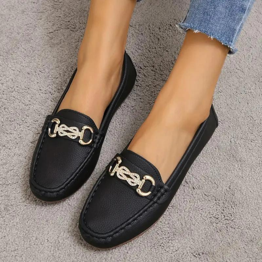 Schwarze Damen-Loafer mit goldener Kettendekoration, getragen auf grauem Teppich. Modische, elegante Schuhe, ideal für Freizeit und Büro.