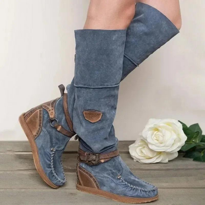 Blaue Damenstiefel aus Wildleder mit braunen Akzenten und Schnallen, auf Holzboden vor weißer Rose. Modische, bequeme Stiefel für Herbst.