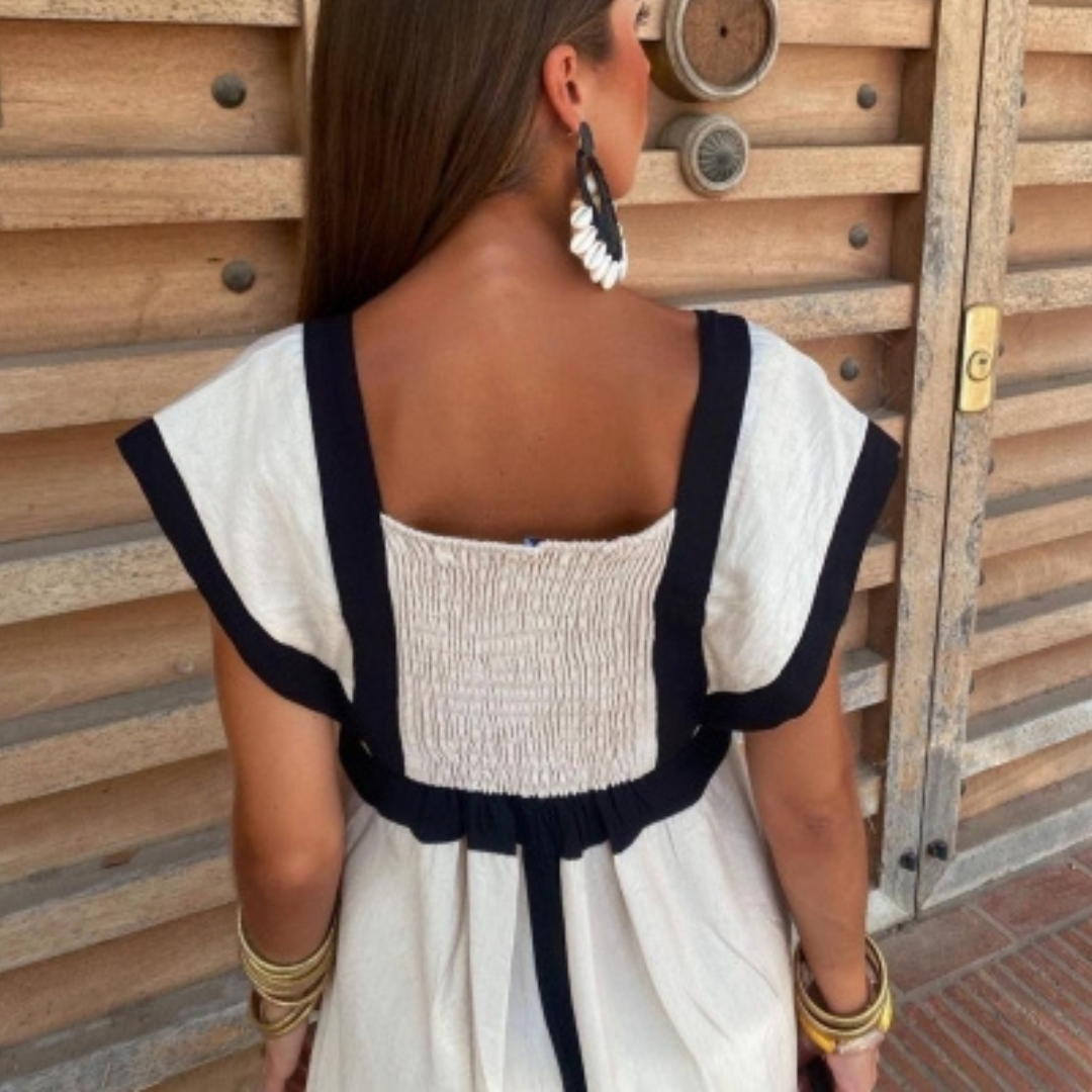 Frau in weißem Sommerkleid mit schwarzen Details, zeigt Rückenansicht. Holzverkleidung im Hintergrund. Mode, Sommerkleid, Damenmode, Accessoires.