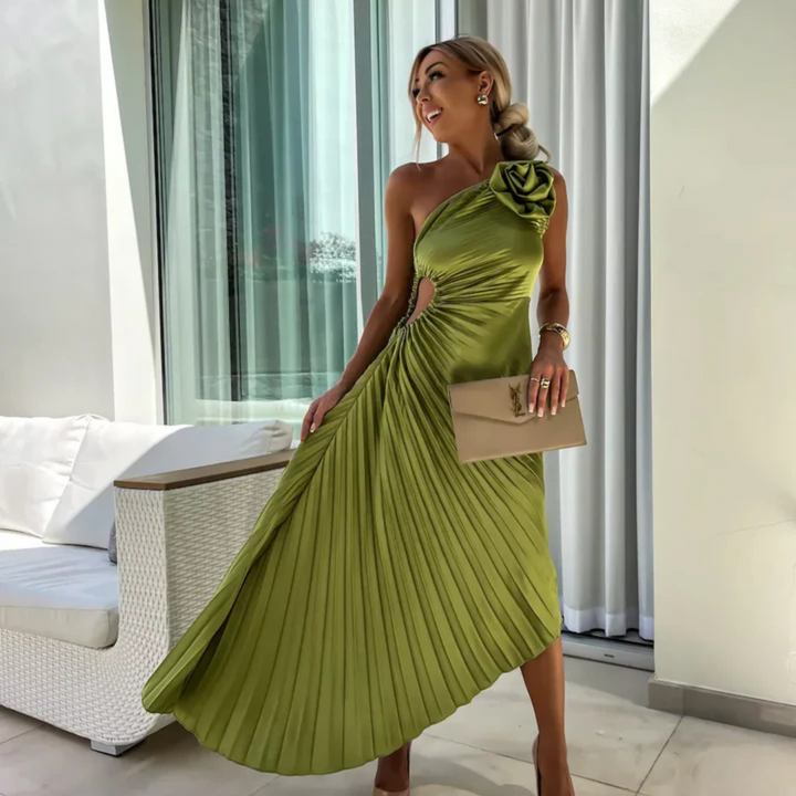 Frau in eleganter, einseitiger, grüner Abendkleid mit Plissee-Design, hält eine beige Clutch, steht vor einem modernen, lichtdurchfluteten Raum.