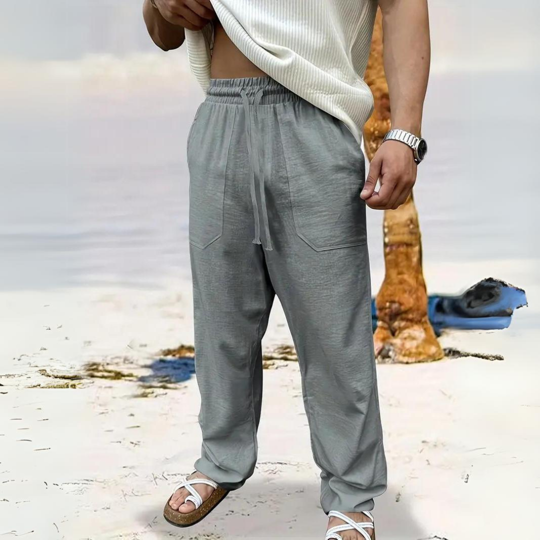 Mann in grauer Leinenhose und weißem Oberteil am Strand, trägt Sandalen. Sommermode, lässiger Stil, Strandkleidung, Herrenbekleidung.