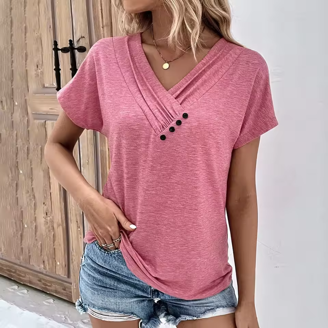 Frau in rosa V-Ausschnitt-T-Shirt mit Knöpfen, kombiniert mit blauen Jeansshorts, steht vor Holztür. Sommermode, lässiger Stil.