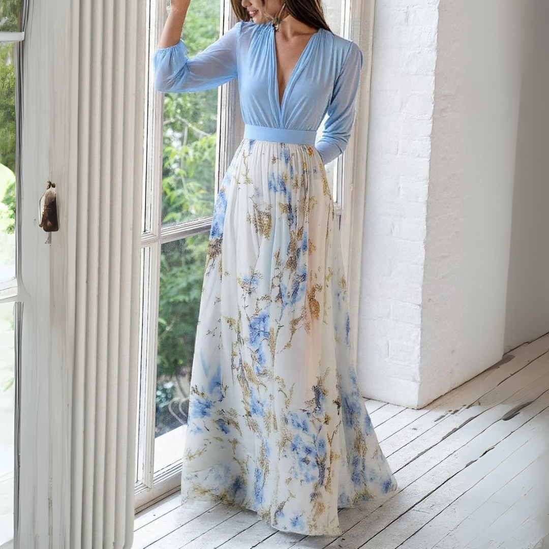 Elegantes langes Kleid mit blauem Oberteil und floralem Rock, stehend vor einem Fenster. Perfekt für Sommermode und Abendveranstaltungen.