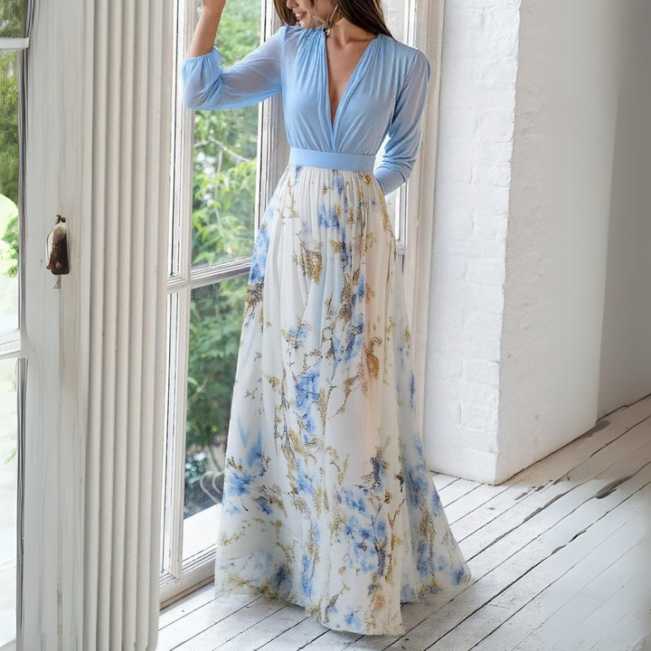 Elegantes langes Kleid mit blauem Oberteil und floralem Rock, stehend vor einem Fenster. Perfekt für Sommermode und Abendveranstaltungen.
